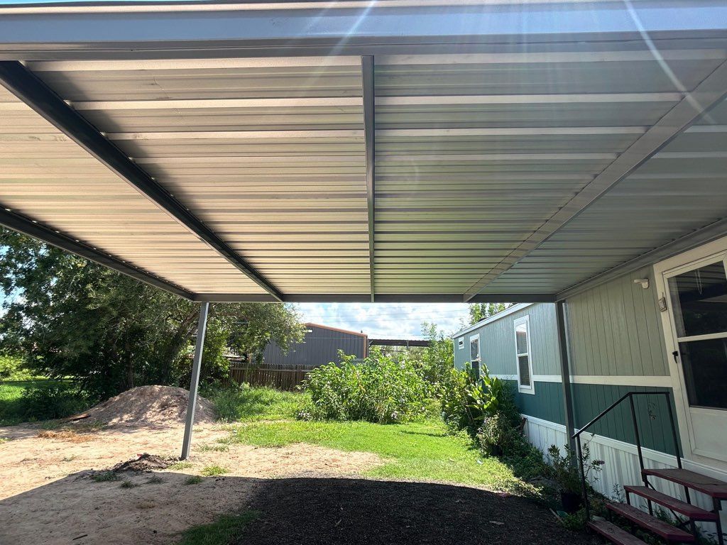 Covered carport beside a white house, with dirt yard, green grass, and trees in the background
