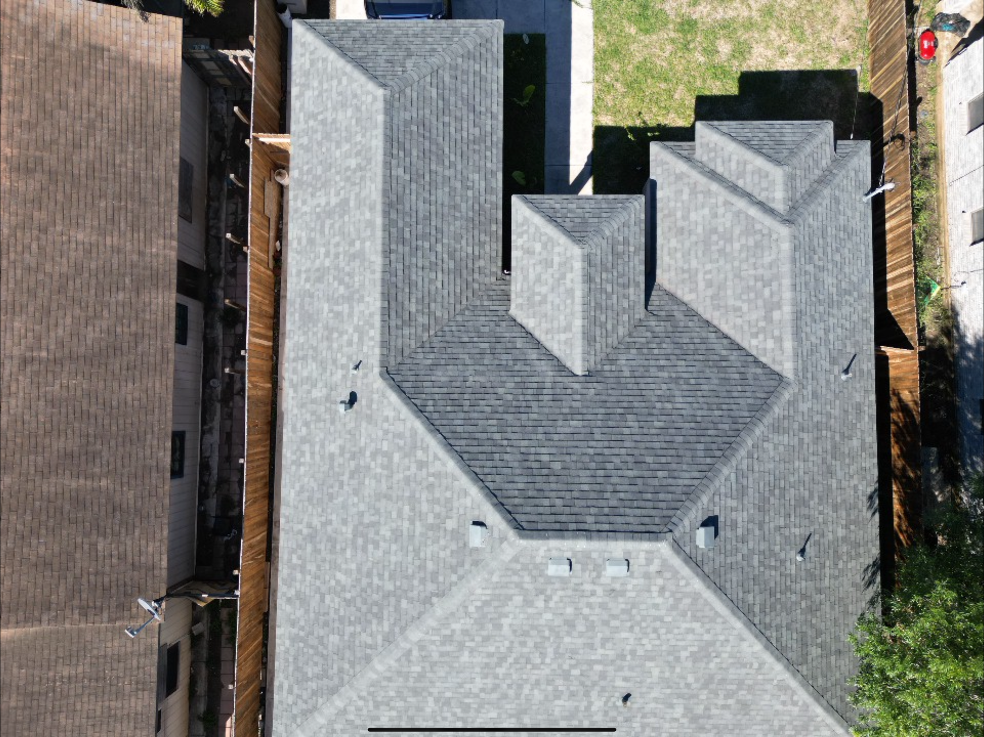 Aerial view of a gray shingled roof with multiple gables and chimneys, between neighboring houses.