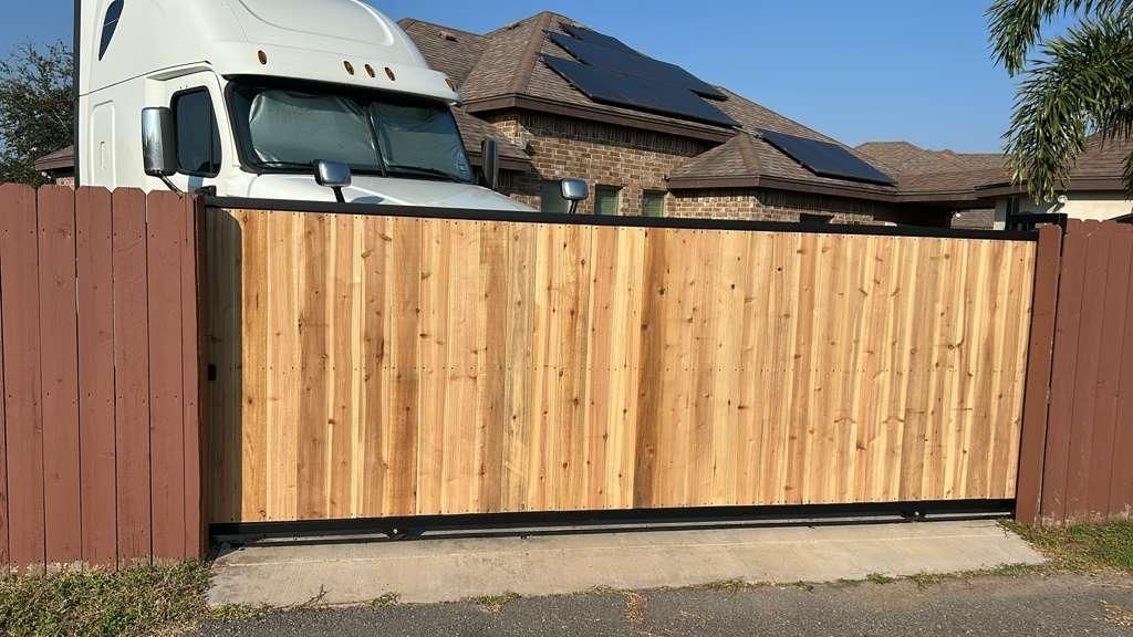 Wooden driveway gate between brown fence sections, with a white truck and house roof visible behind it.