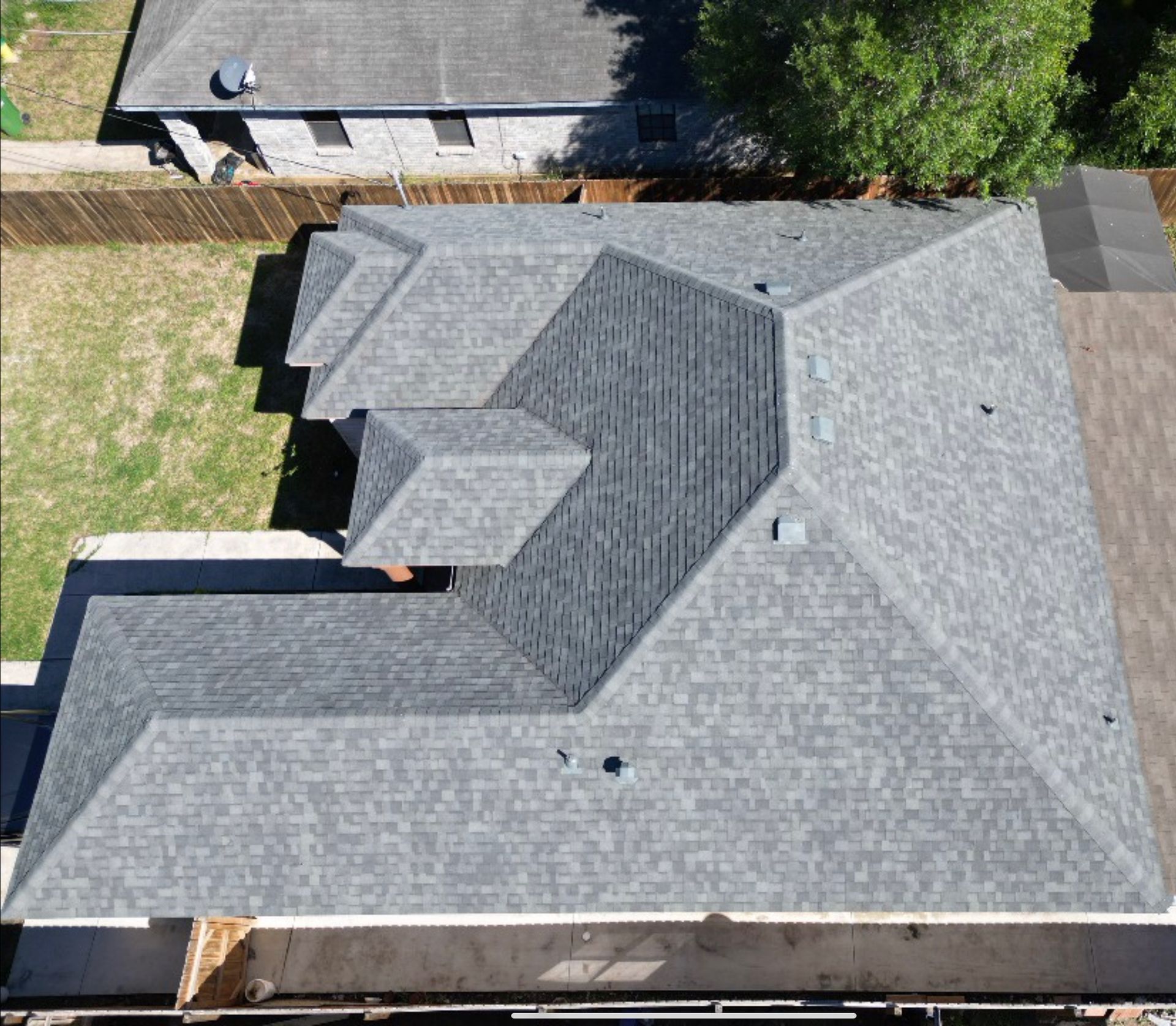 Aerial view of a gray shingled house roof with multiple gables and vents beside a grassy yard.