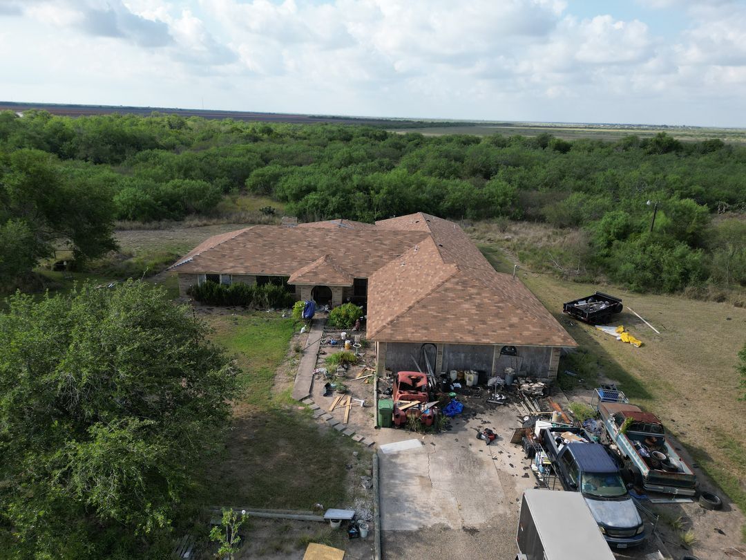 Aerial view of a rural house with a tan roof, cars parked outside, surrounded by dense green trees.
