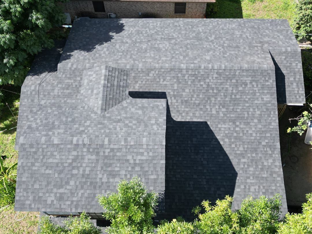 Aerial view of a gray-shingled house roof with multiple gables and surrounding trees.