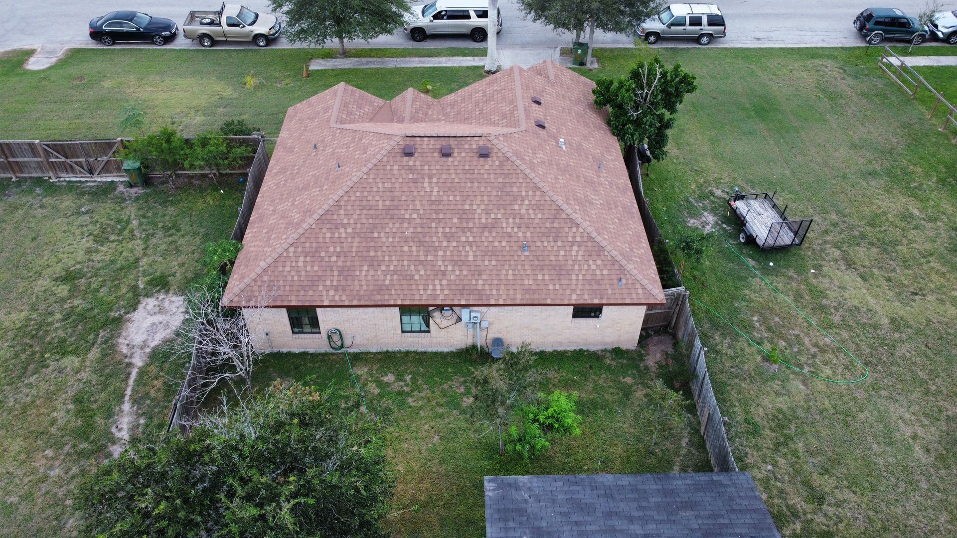 Aerial view of a small tan house with a red roof, fenced yard, and parked cars along the street.