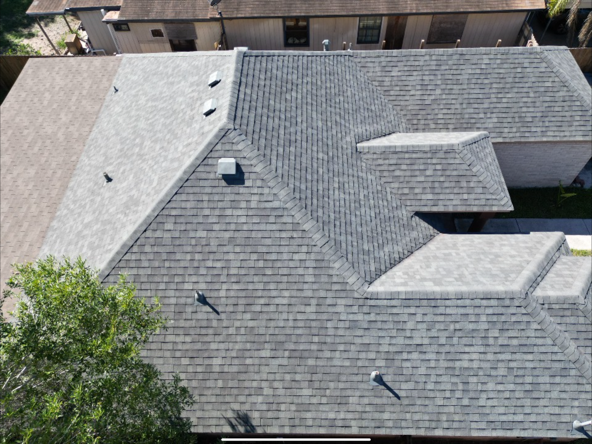 Aerial view of a gray shingled rooftop with vents and skylights beside trees and neighboring buildings
