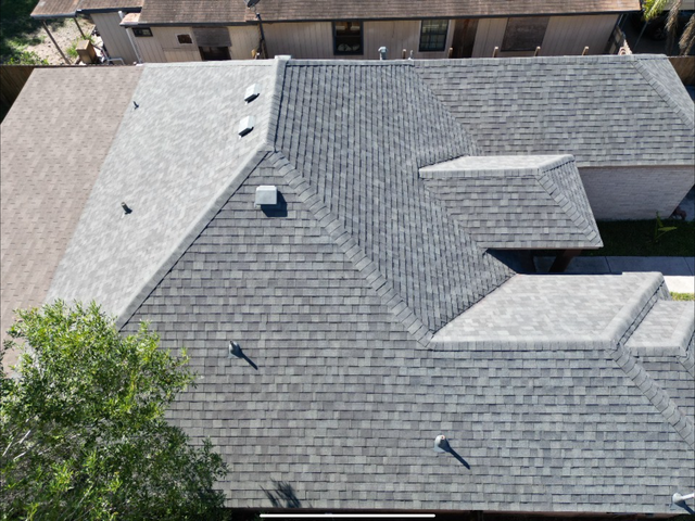Aerial view of a gray shingled rooftop with vents and skylights beside trees and neighboring buildings