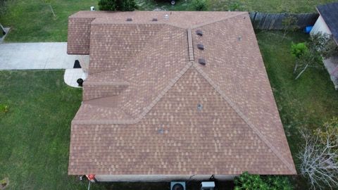 Aerial view of a tan shingled house roof with dormer, vents, driveway, and green yard.