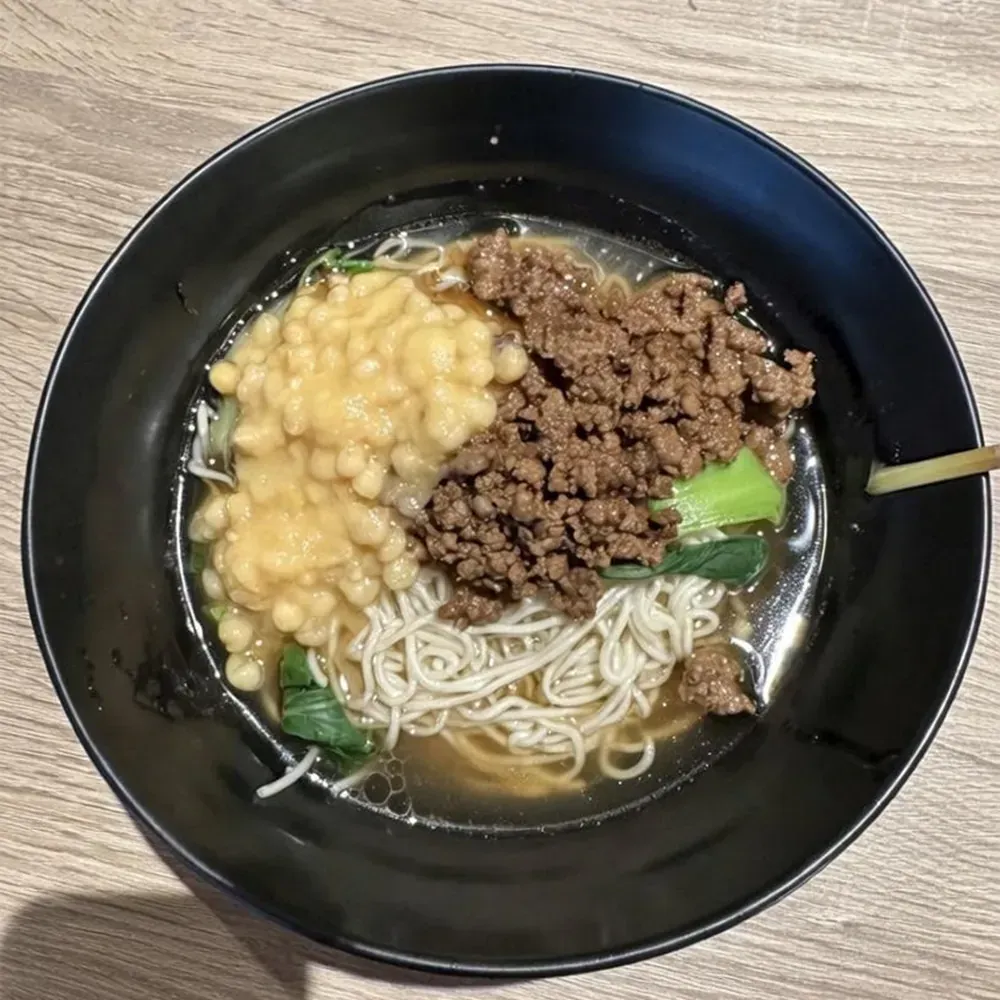 Black bowl of noodle soup topped with minced meat, corn, greens, and chopsticks on a wooden table