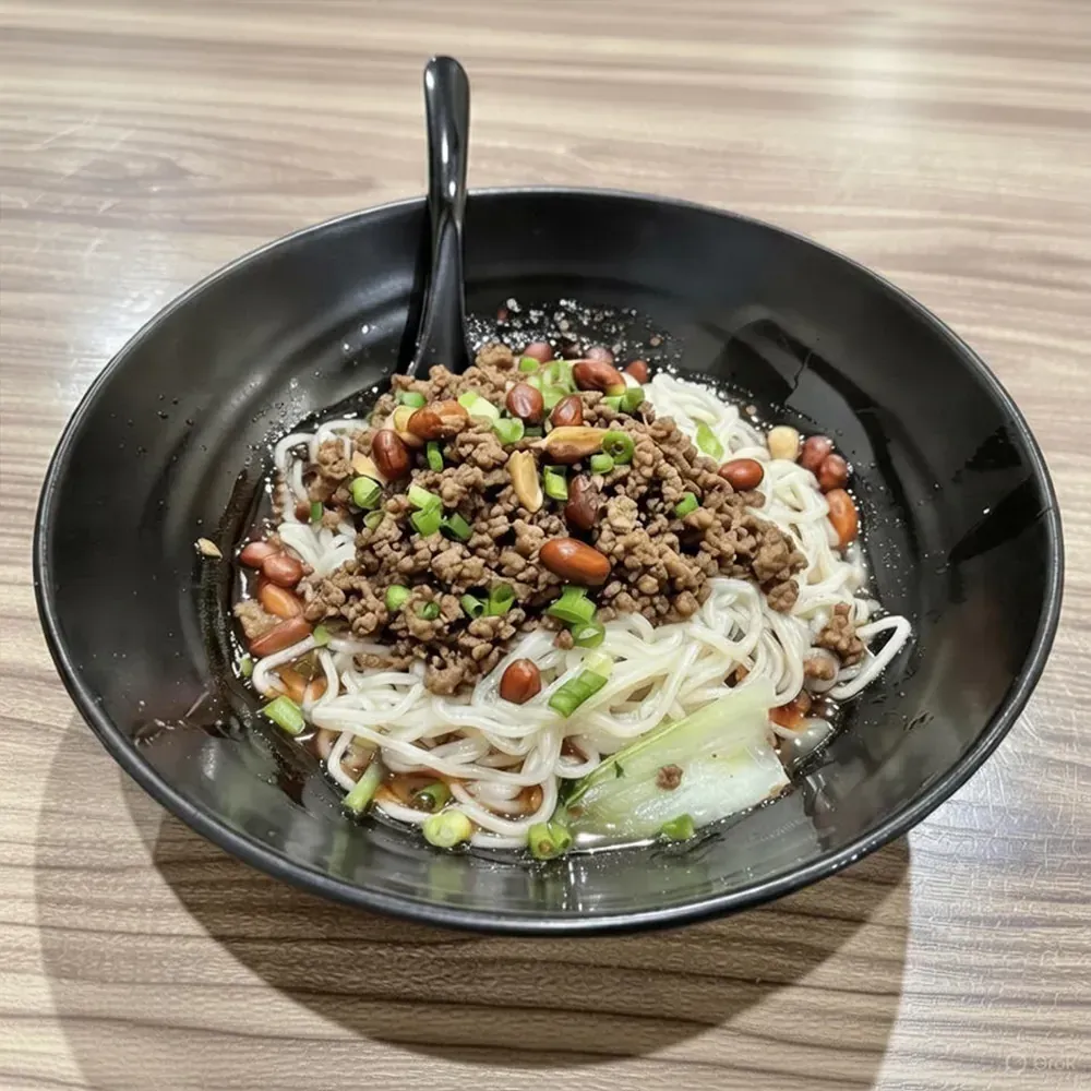 Black bowl of noodle soup topped with ground meat, peanuts, and scallions, with a spoon on a wooden table