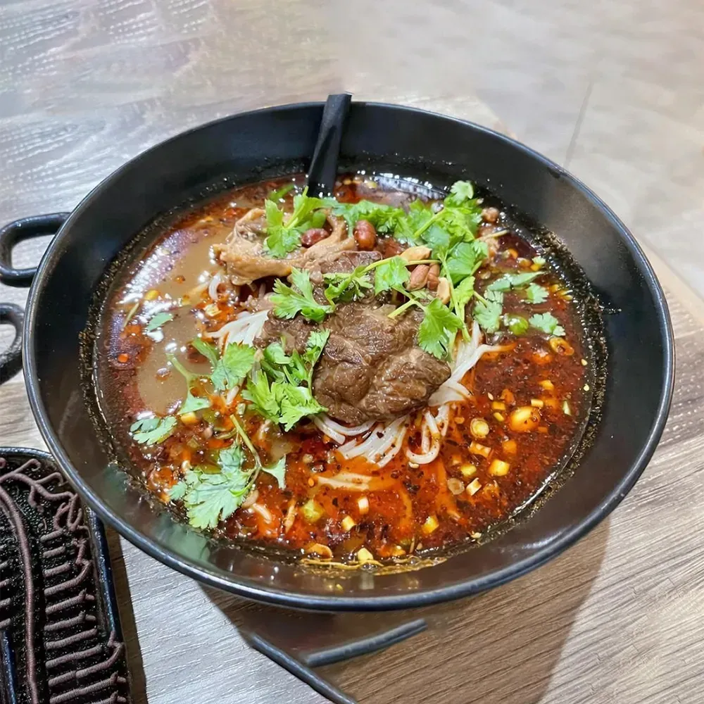 Spicy noodle soup with beef, cilantro, and chili oil in a black bowl on a wooden table