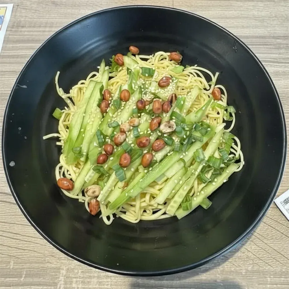 Black bowl of noodles topped with sliced cucumbers, peanuts, and green sauce on a wooden table