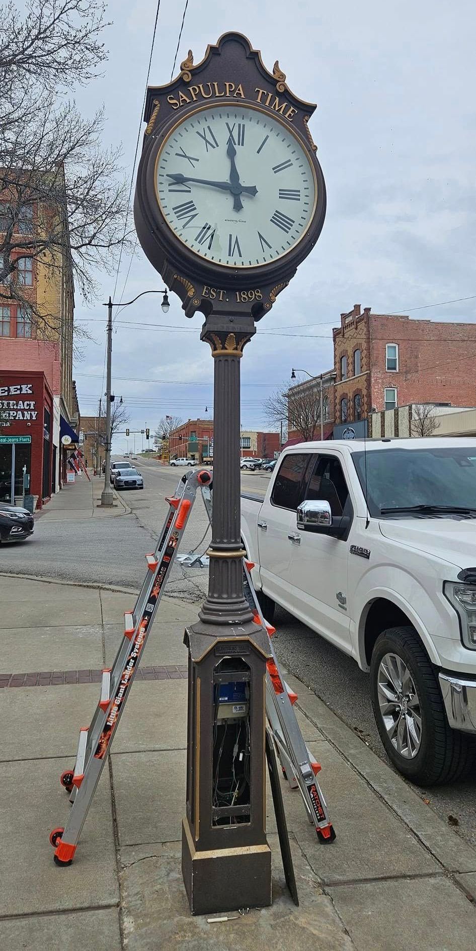 A clock is sitting on the side of a street next to a white truck.