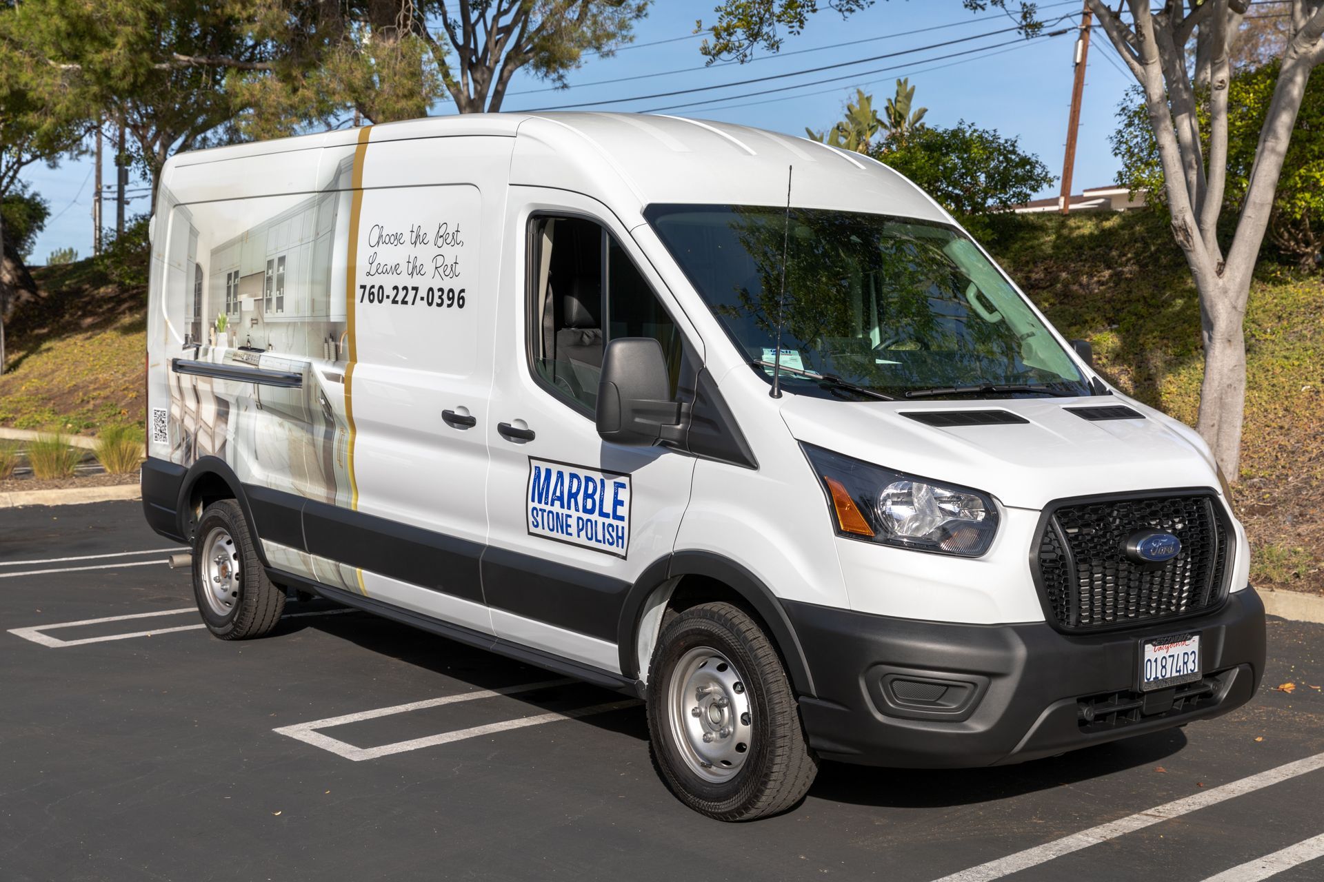 A white Ford Transit commercial van with branded decals parked in an outdoor parking lot under a clear blue sky.