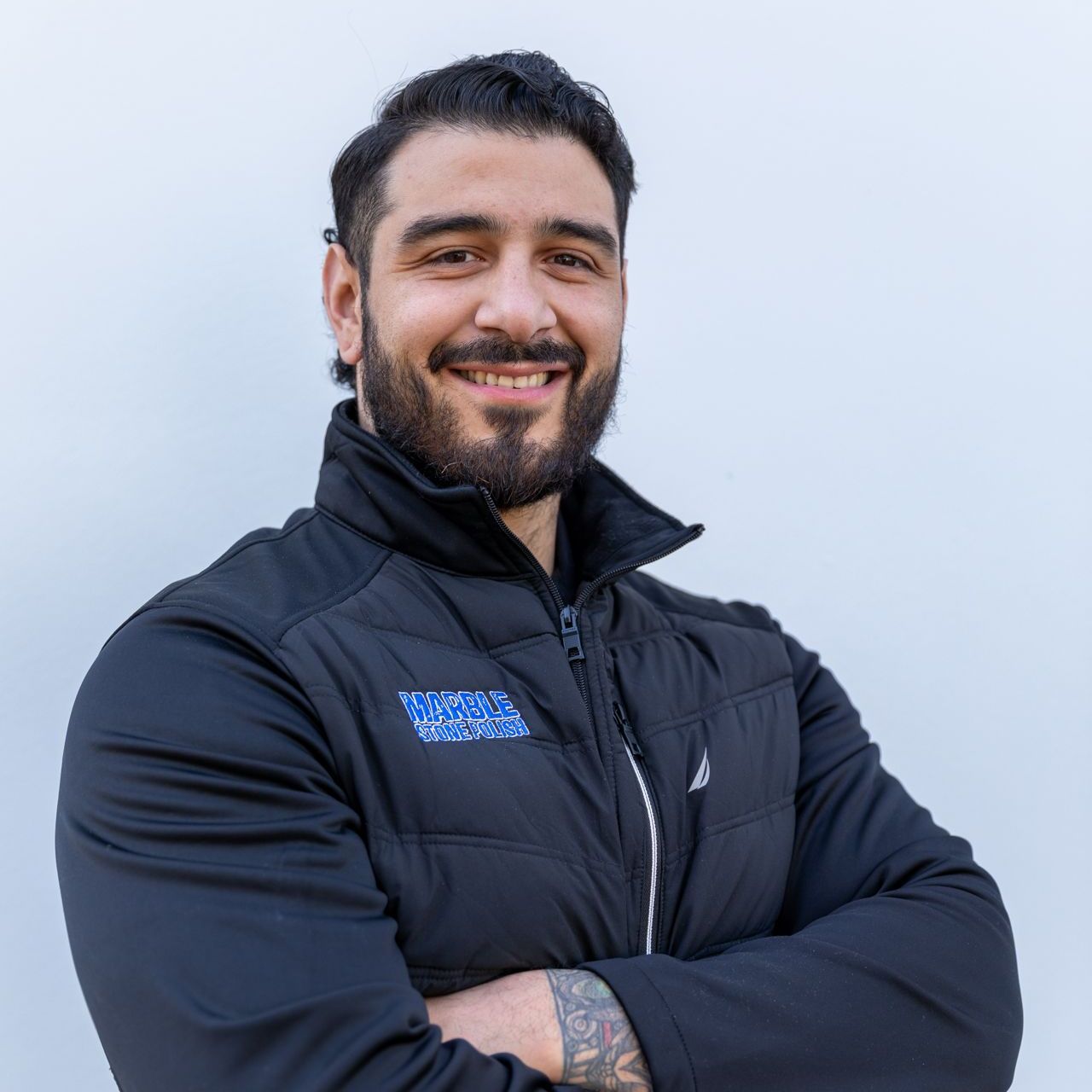 A person with a beard and dark hair smiles with arms crossed, wearing a black jacket against a plain light background.