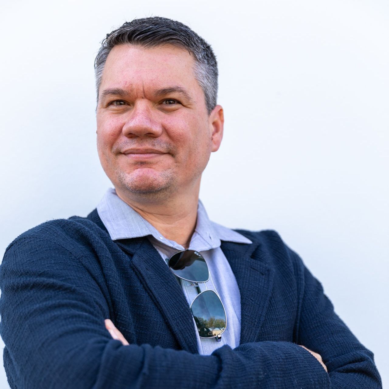 A person with short, dark hair wearing a blue blazer and light shirt, standing with arms crossed against a white wall.