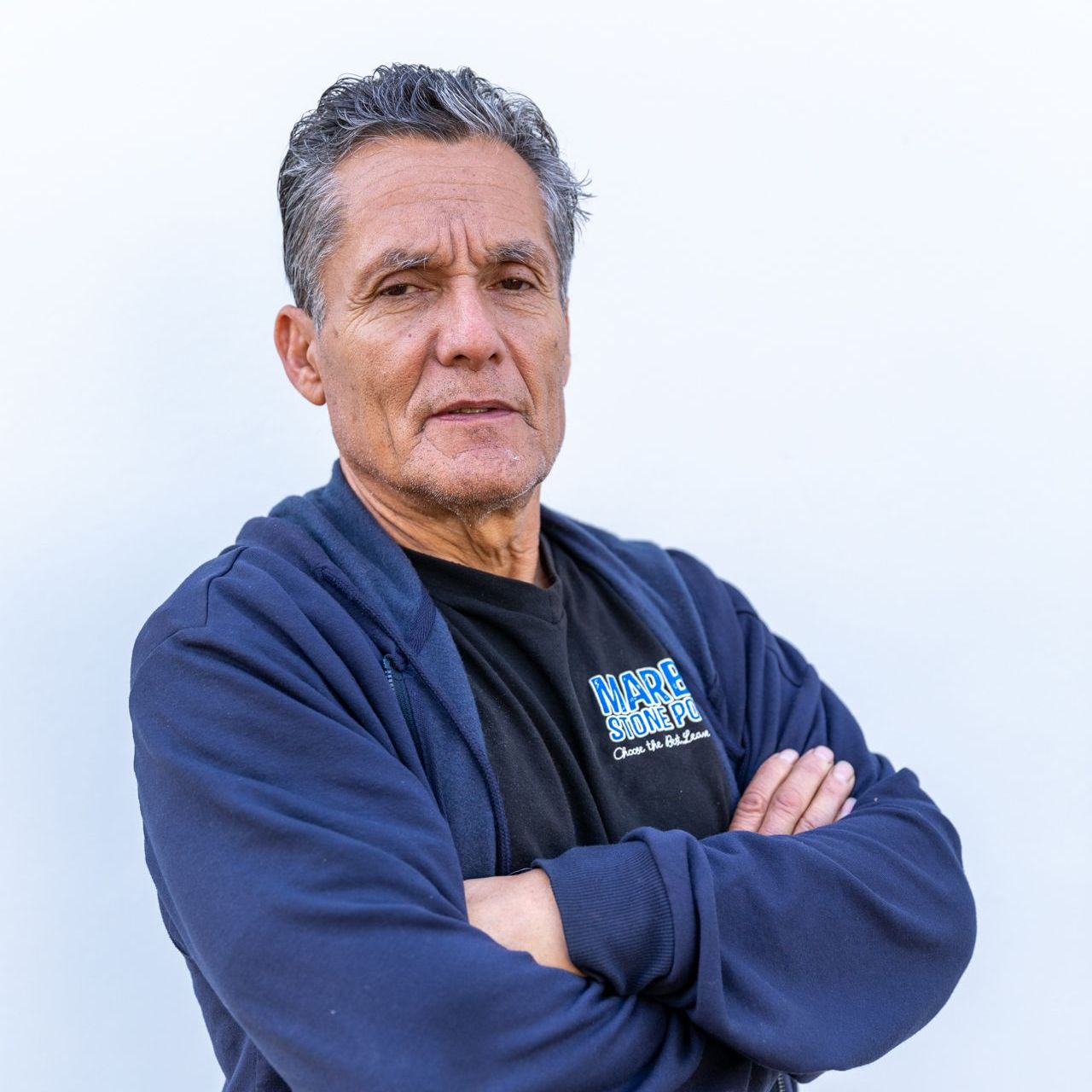 A man with salt-and-pepper hair, wearing a dark blue hoodie and black t-shirt, stands with arms crossed against a wall.