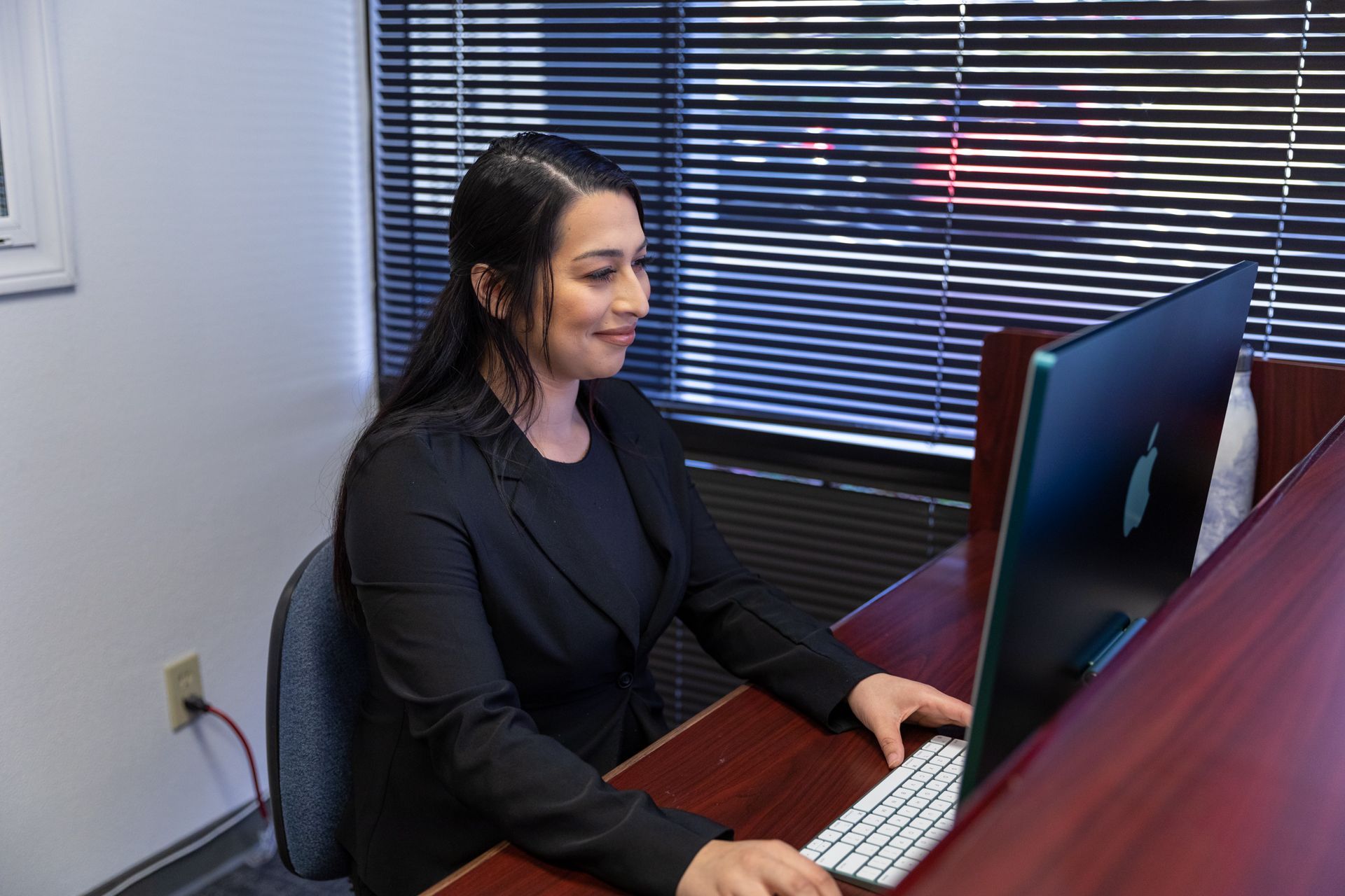 A person in a black blazer smiling while working at a computer in an office.