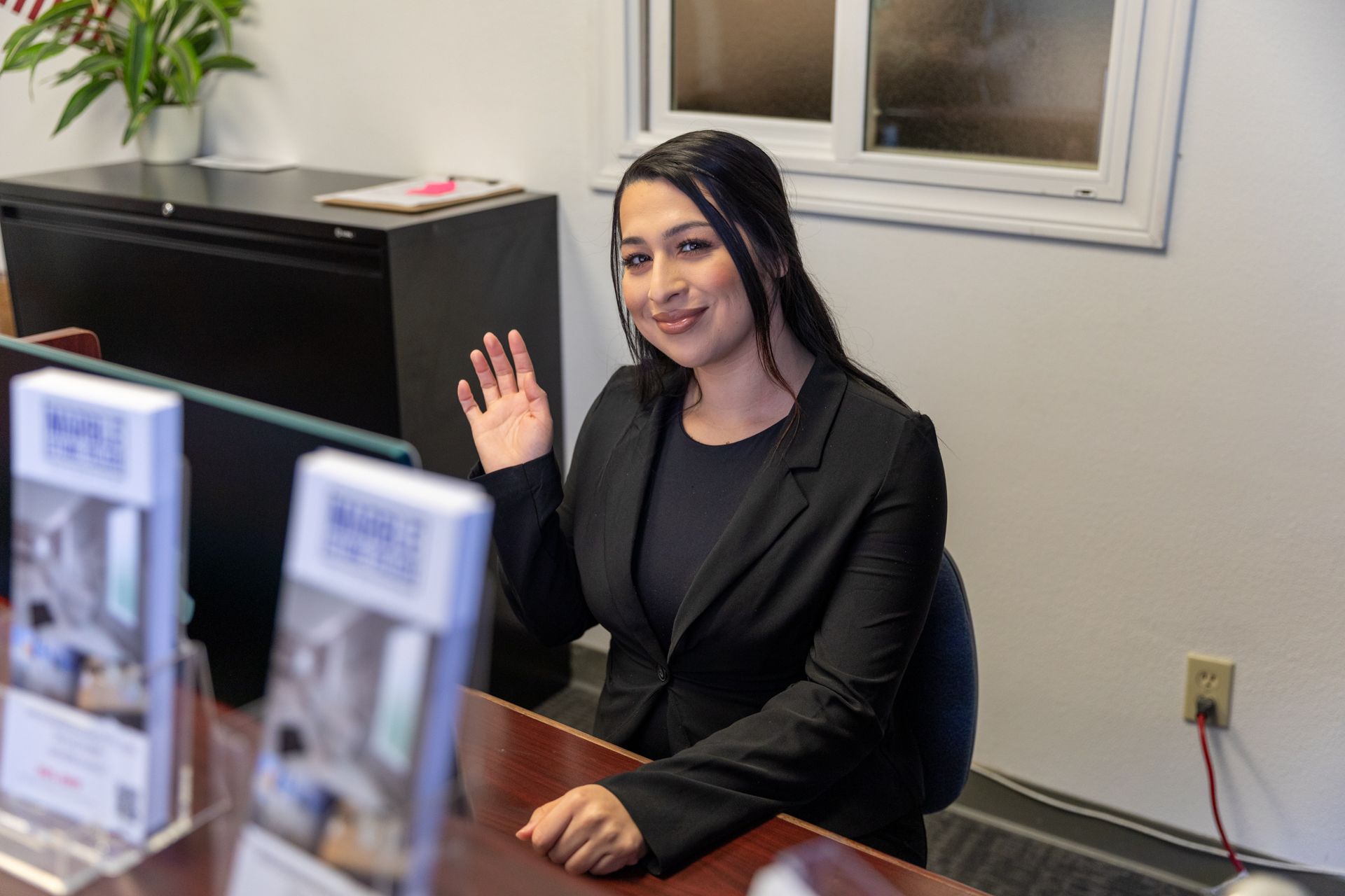 A smiling person in a dark blazer waves from a desk in an office with brochures in the foreground.