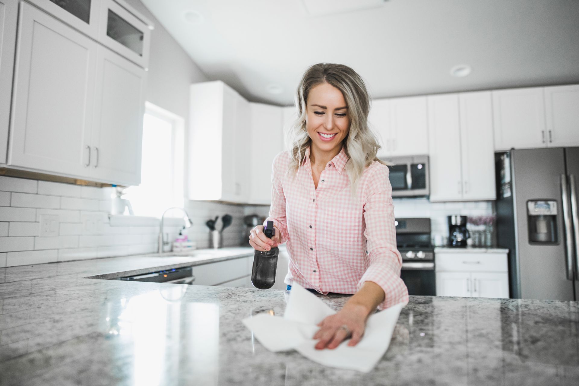 Woman cleaning a white countertop in a bright kitchen with a spray bottle and cloth.