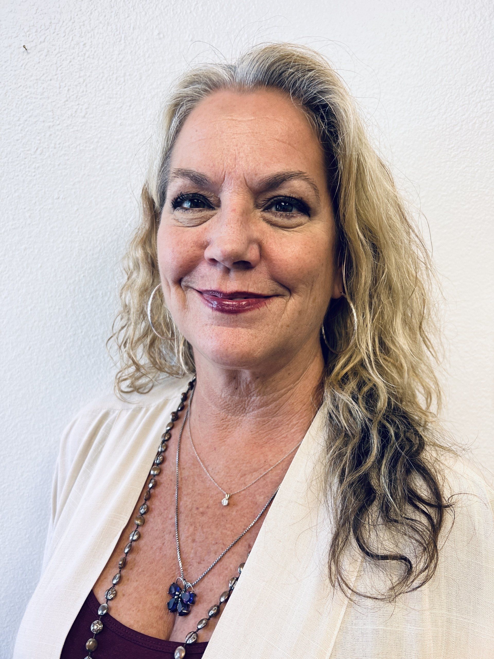 A person with long, curly light hair and a gentle smile, wearing a white cardigan and layered necklaces against a wall.