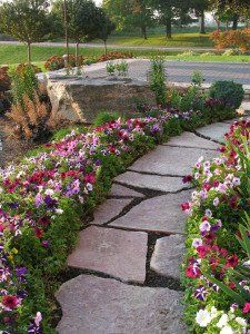 A stone path curves through a garden filled with vibrant, multi-colored flowers leading to a large rock and a paved area.