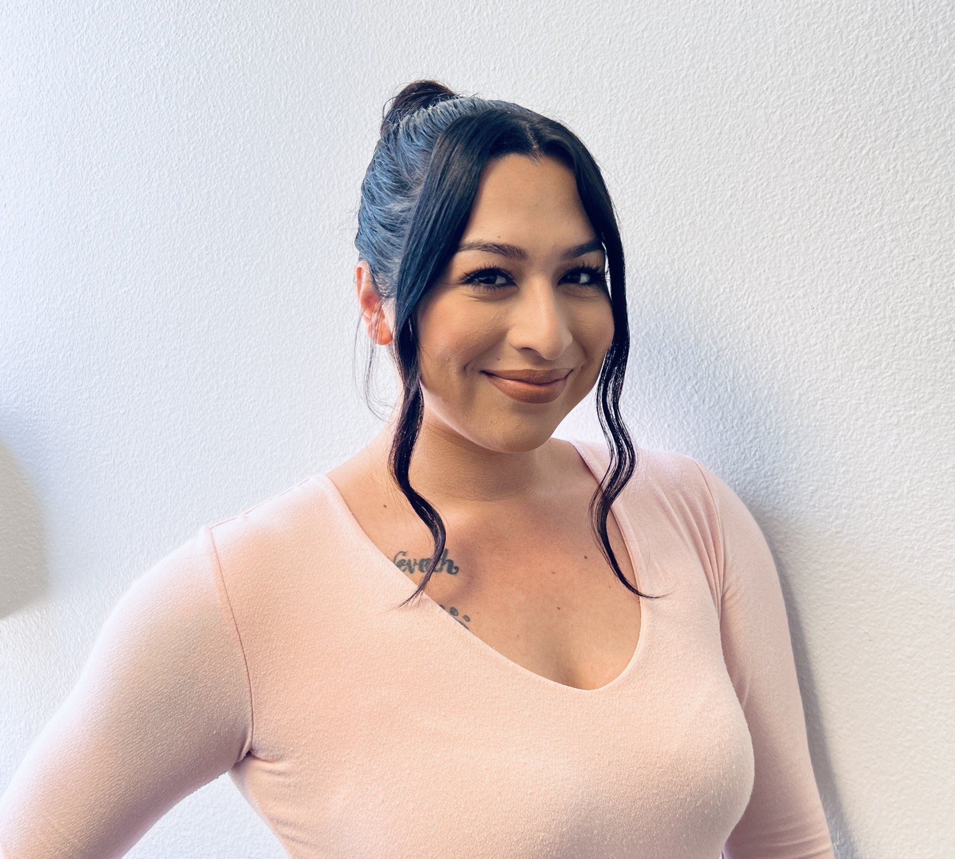 A person with dark hair styled in an updo and loose strands smiles while wearing a light pink long-sleeved top.
