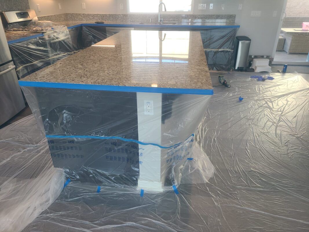 A kitchen island with tan granite countertops covered in protective plastic and blue painter's tape for renovations.