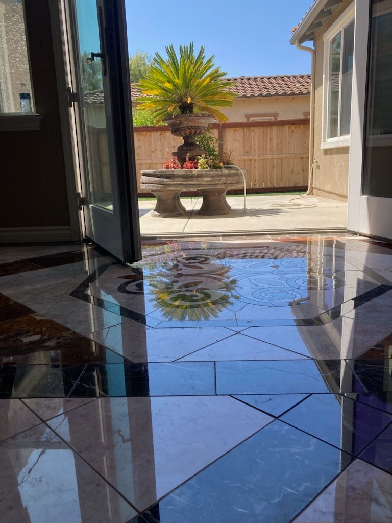 A polished, reflective floor with a geometric pattern mirrors a sunlit patio featuring a fountain and palm tree.