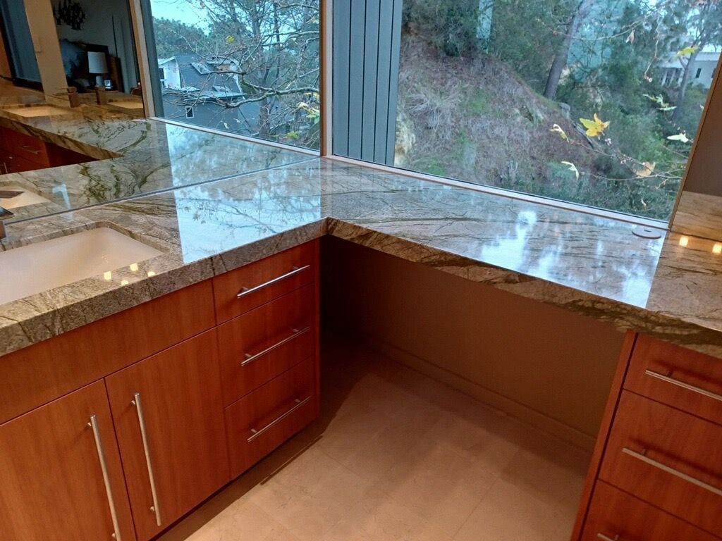 A high-angle view of a bathroom vanity with granite countertops, wooden cabinets, and a large window looking outdoors.