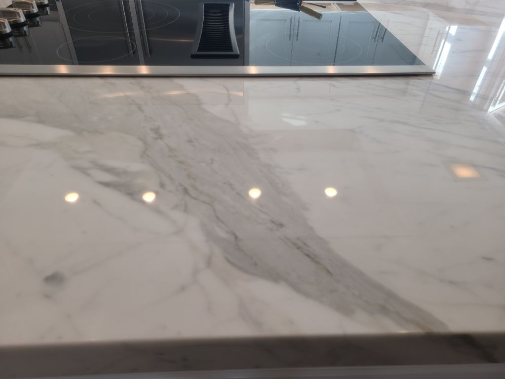 A close-up of a white marble countertop with grey veining, featuring a black stovetop in the background.
