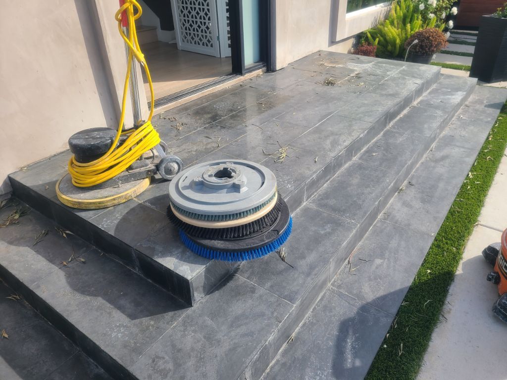 A floor buffer and interchangeable circular cleaning brushes sit on a wet, dark stone porch outside a building.