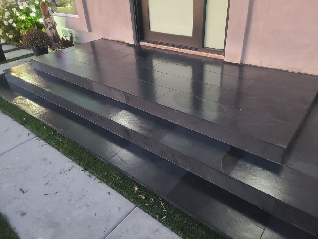 A set of two dark, glossy tiled steps leading to a house entrance, bordered by a patch of green grass.