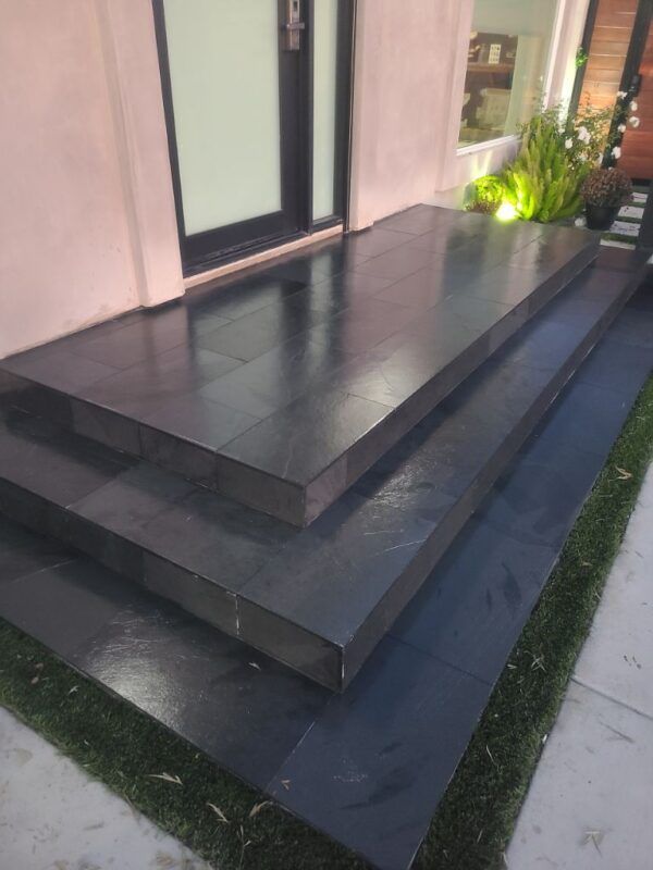 Two dark, tiled steps leading to a building entrance, with a small patch of artificial grass along the bottom edge.