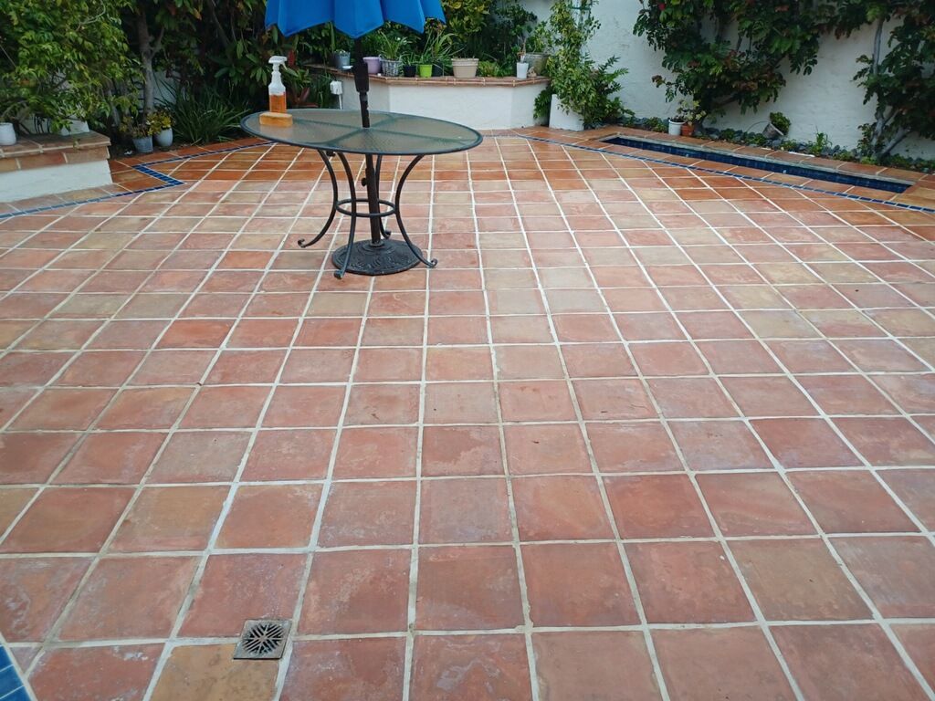 A patio paved with square terracotta tiles features a black metal table, a blue umbrella, and a small built-in drain.