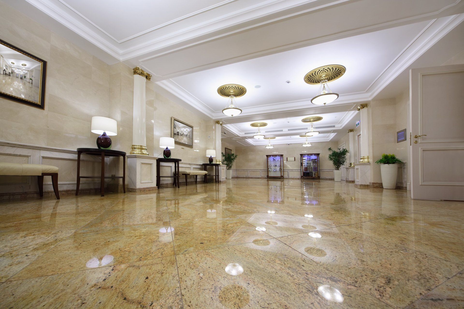 A wide, polished marble-floored lobby with beige walls, ornamental ceiling lights, and wooden accent tables.