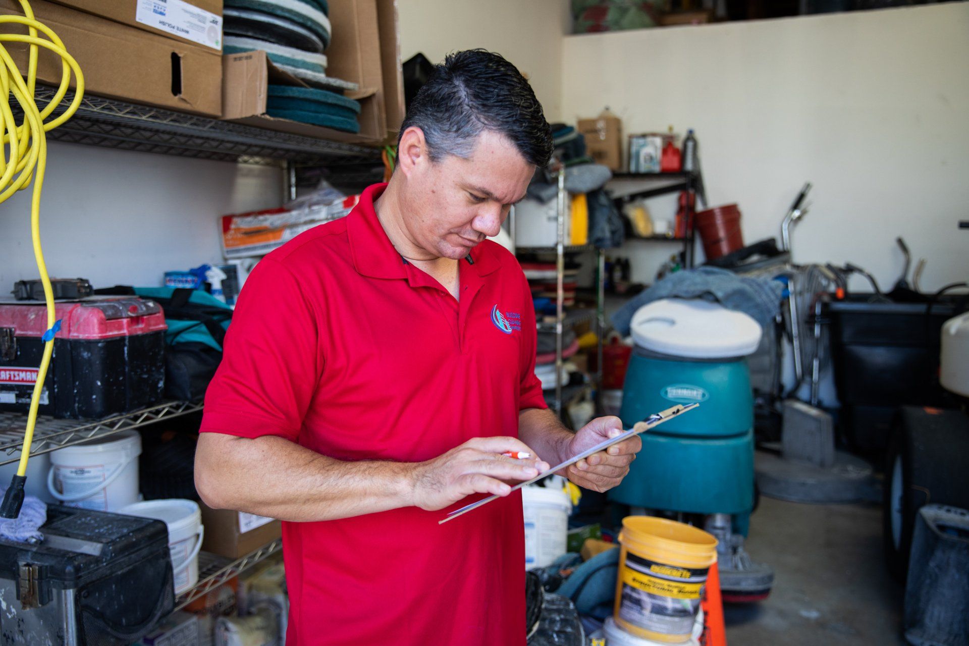 A person in a red polo shirt reviews a clipboard in a storage room filled with cleaning supplies and equipment.