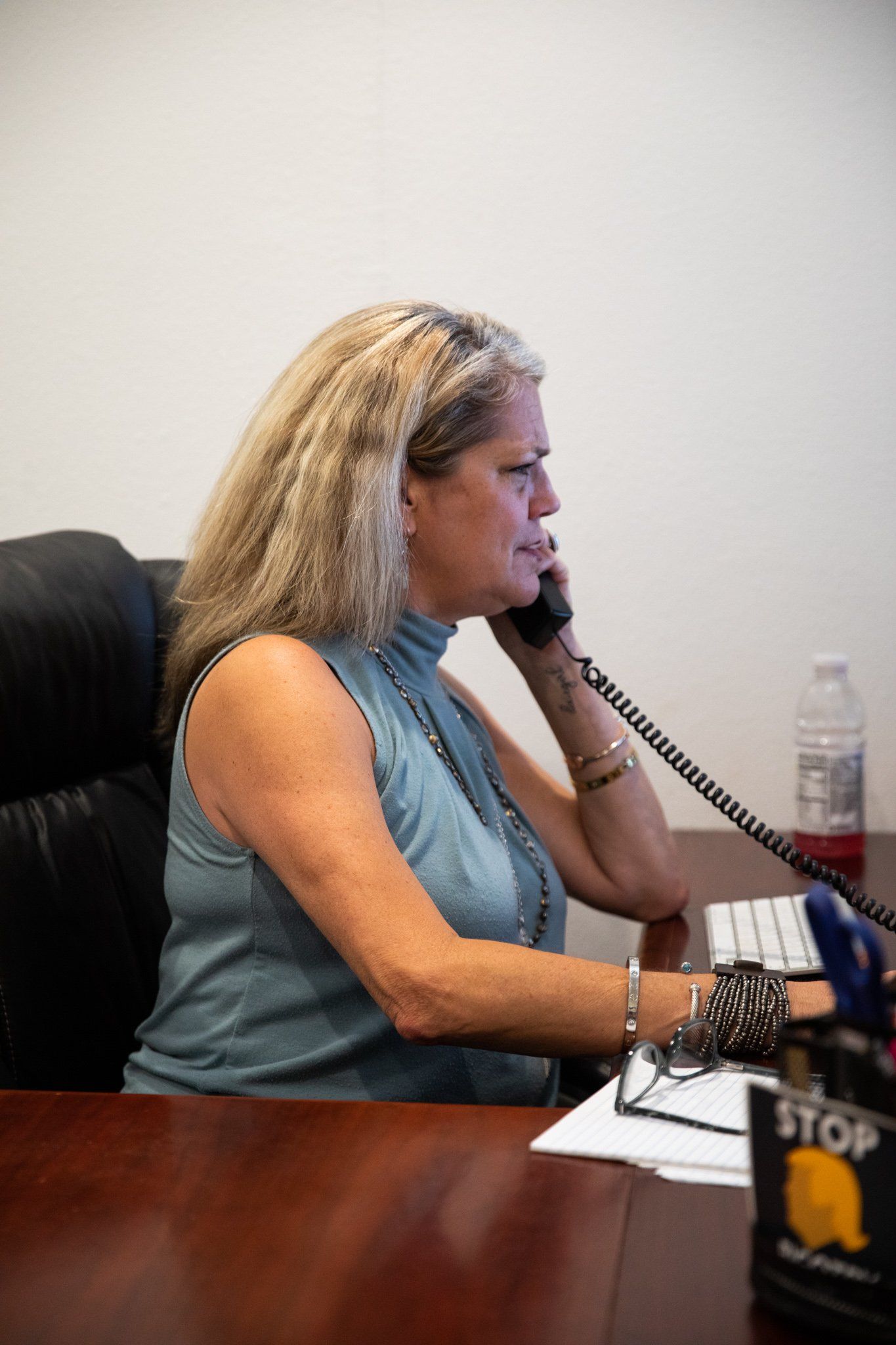A person with wavy blonde hair wears a light blue sleeveless top and talks on a corded phone while seated at a desk.