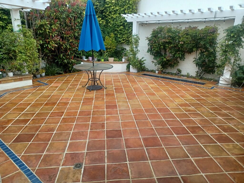 A patio with red-brown square tiles, a round table with a blue umbrella, and green foliage against a white wall.