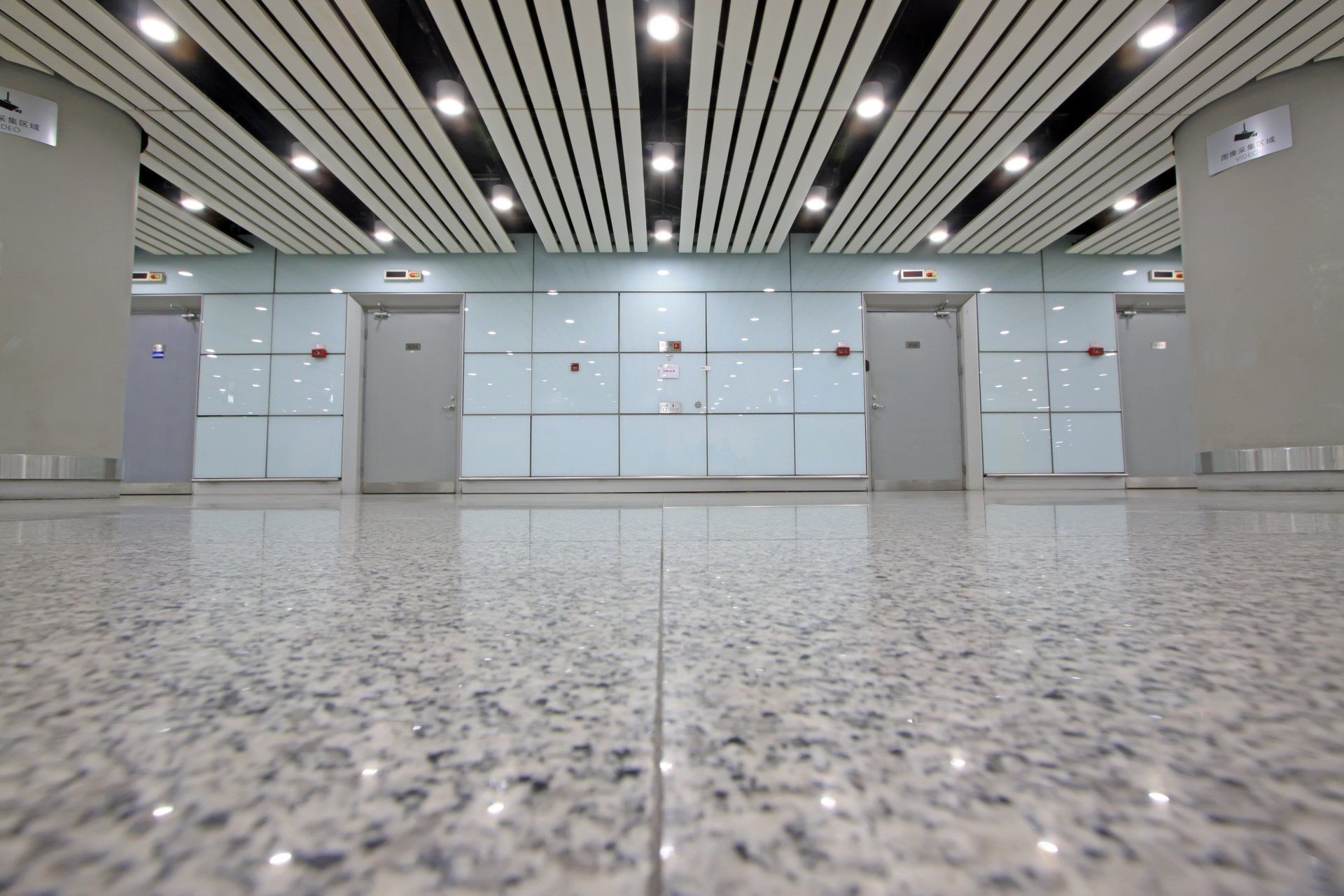 A low-angle view of a clean, brightly lit hallway with gray tiled walls, several doors, and a polished stone floor.