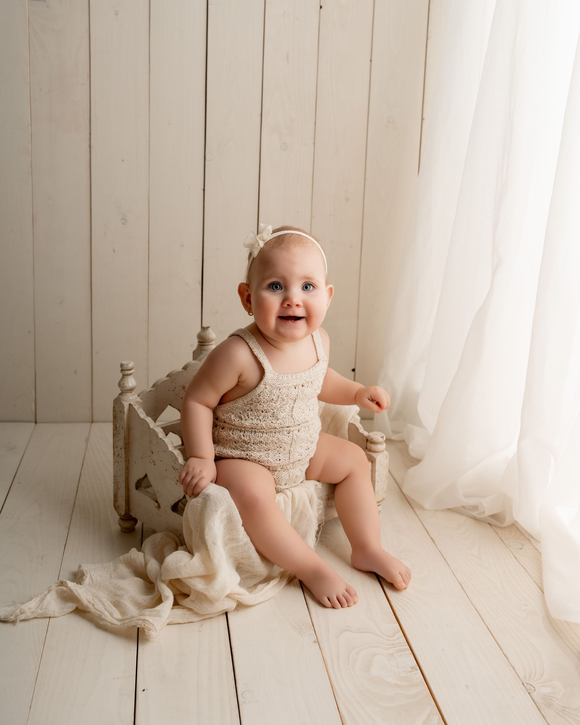 Een lachende baby in een beige gehaakt pakje met hoofbandje zit in een klein, verweerd wit bedje; lichte, heldere achtergrond.