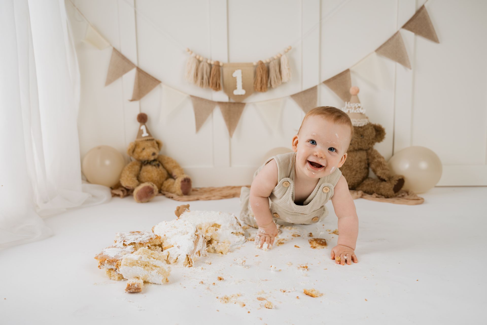 Een peuter kruipt rond in een beige decoratie voor een eerste verjaardag, compleet met teddyberen, slingers en taartkruimels op de vloer.