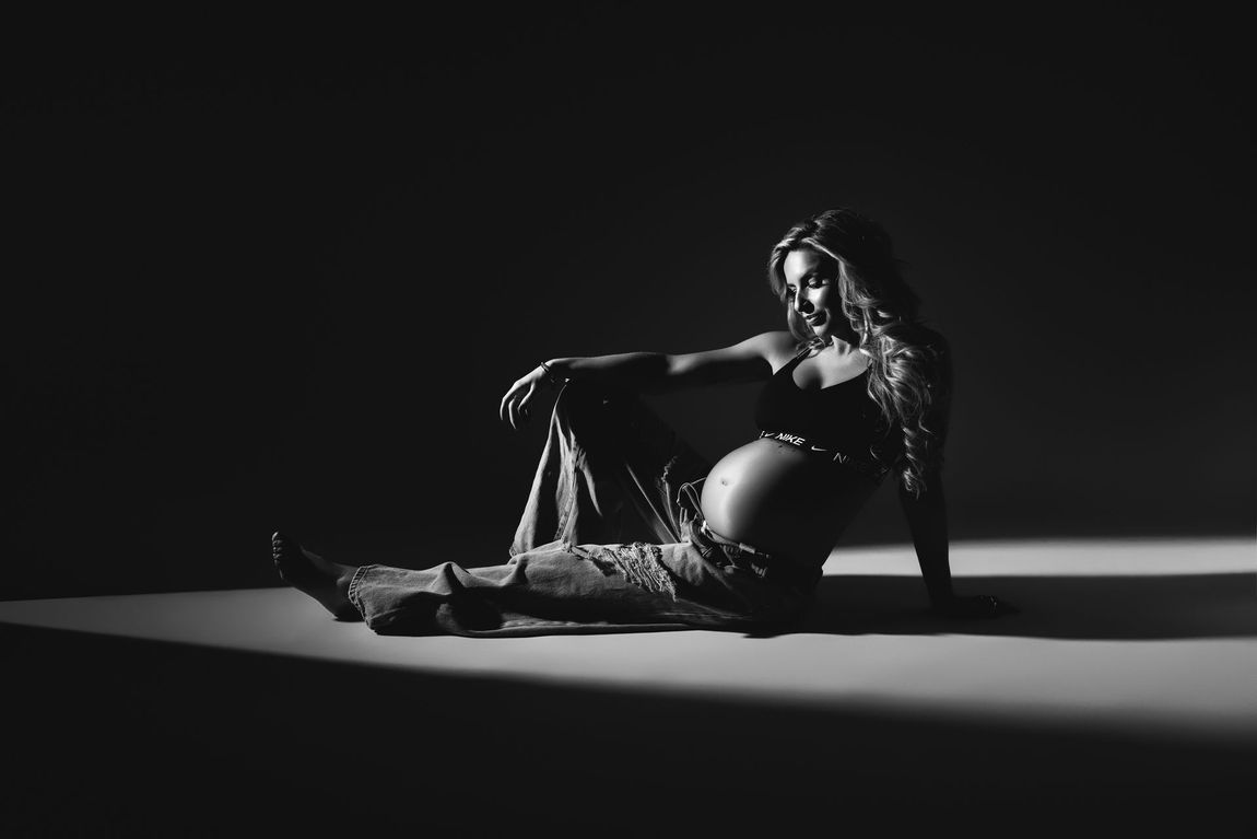 Een zwangere vrouw zit op de grond in een dramatisch zwart-wit studiofoto.