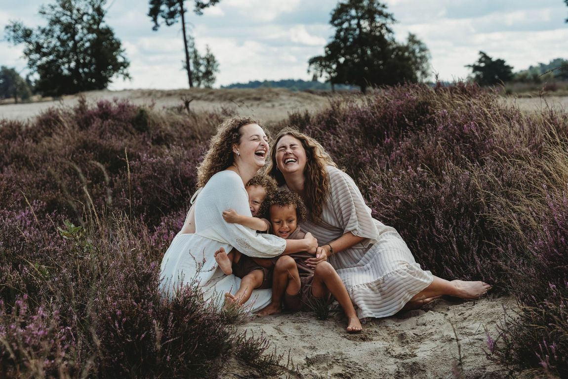 Drie mensen zitten te lachen in een grasveld met hoge struiken op de achtergrond.