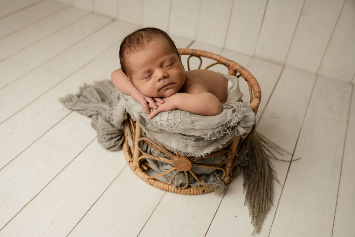 Een slapende baby ligt in een klein rieten mandje op een witte houten vloer, gewikkeld in een grijze deken.