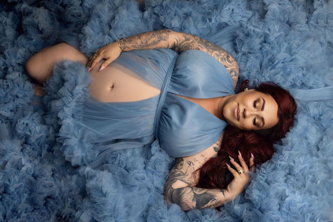 Vrouw in een blauwe jurk ligt op een blauwe achtergrond van imitatiebont, met gesloten ogen en zichtbare tatoeages op haar armen.