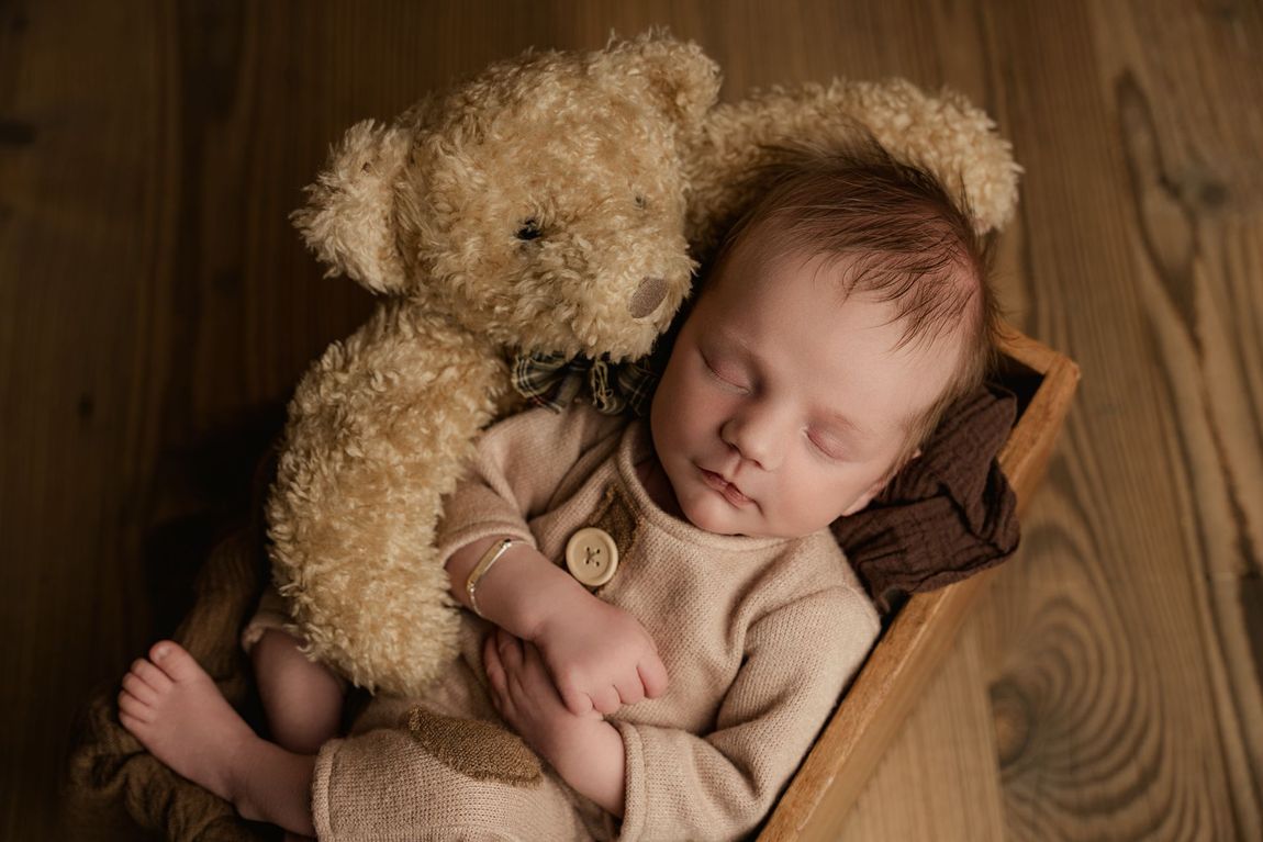 Een slapende baby ligt dicht tegen een teddybeer aan in een houten kist op een houten vloer.