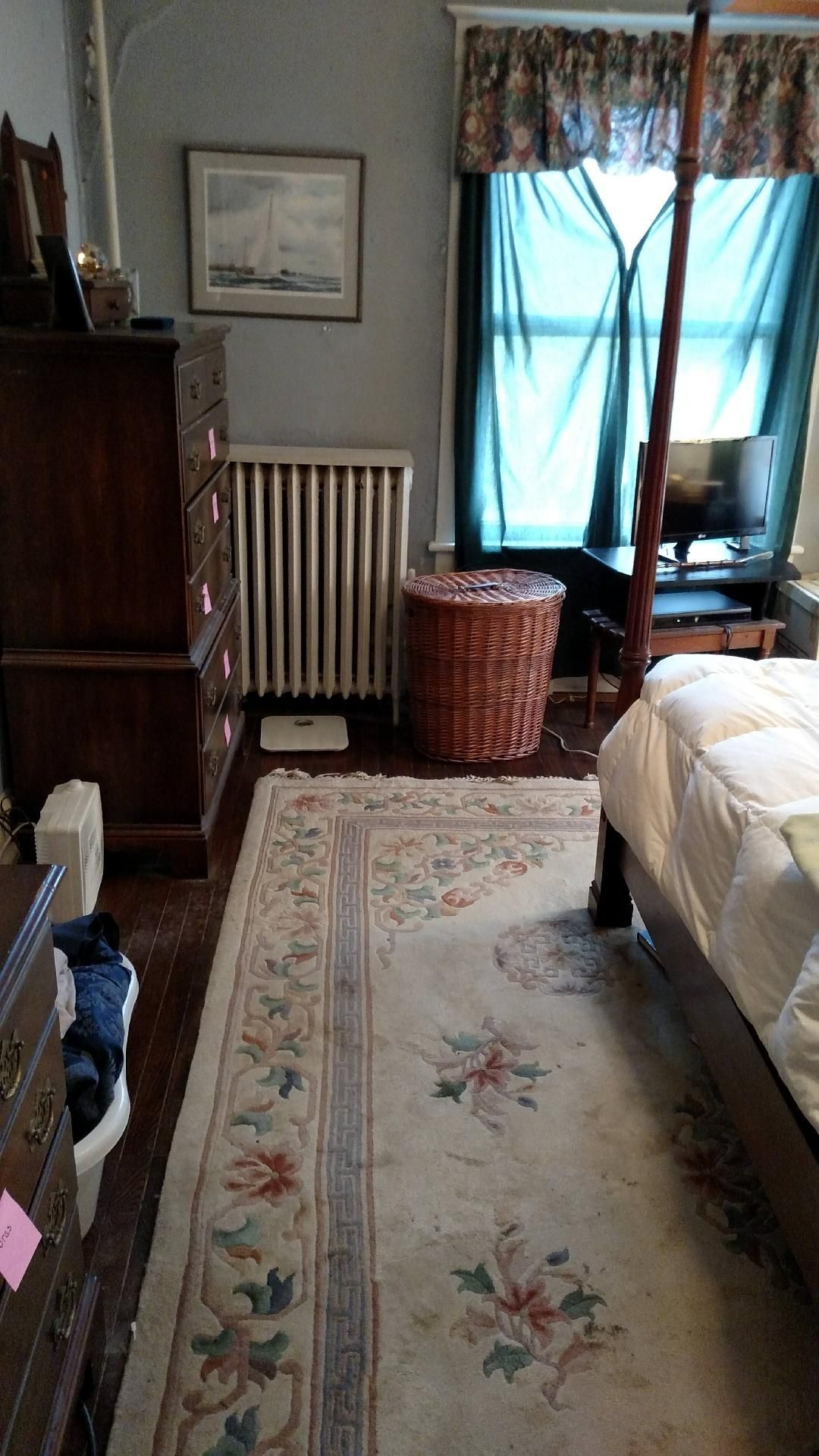 A bedroom with a four poster bed , dresser , rug , radiator and window.