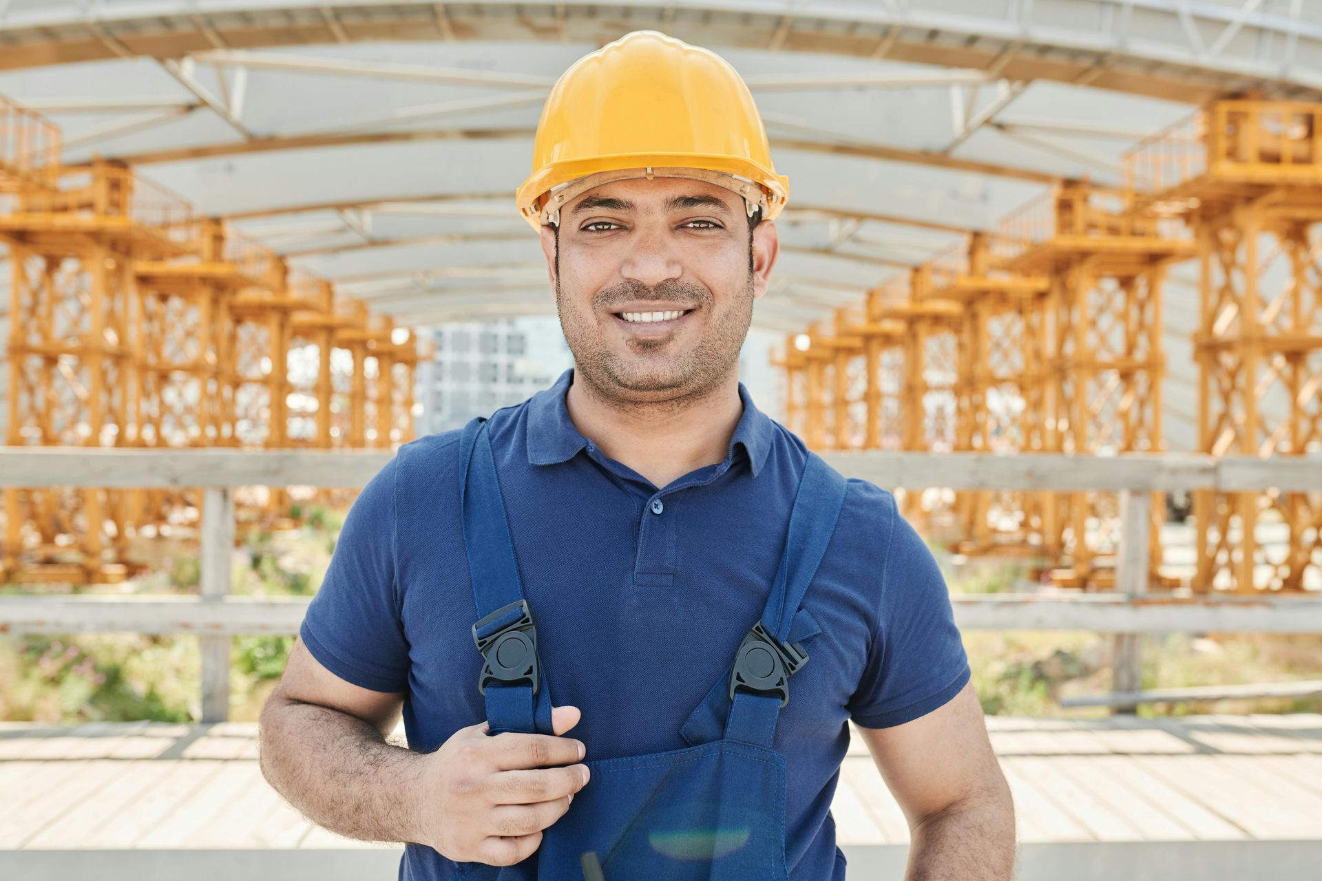 Smiling construction worker wearing a yellow hard hat and blue overalls, in front of construction equipment.