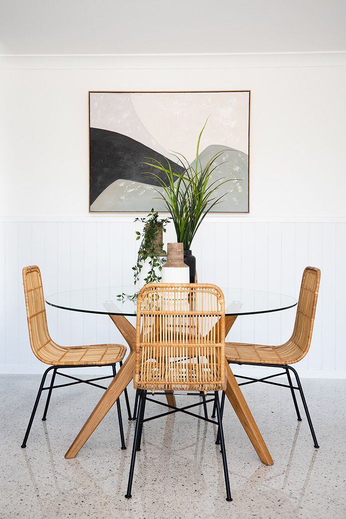 Dining room with round glass table, woven chairs, abstract art, and greenery.