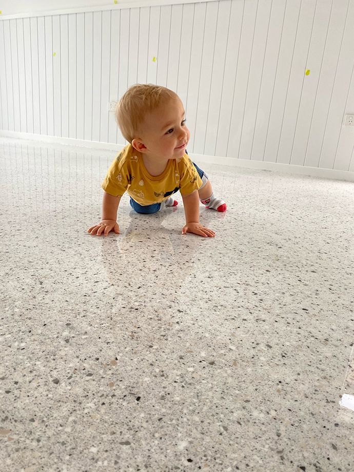 Smiling baby crawling on a speckled, light-colored floor, wearing a yellow shirt and blue pants. White wall in background.