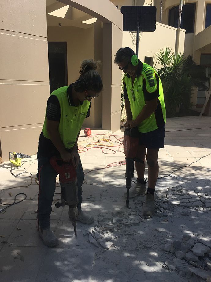Two workers in safety vests break concrete with jackhammers outdoors.